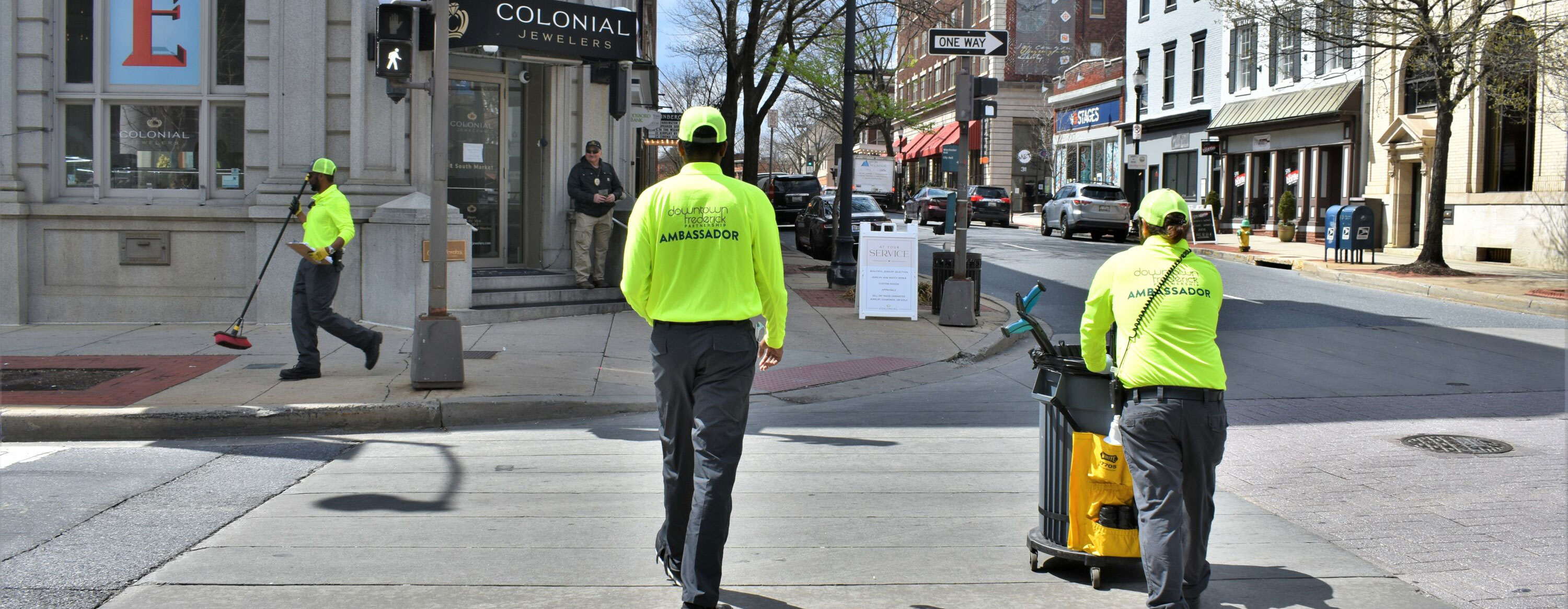 2 people are crossing the street. One of them is pushing a trash can cart. In the background, a 3rd person is walking while carrying a broom.