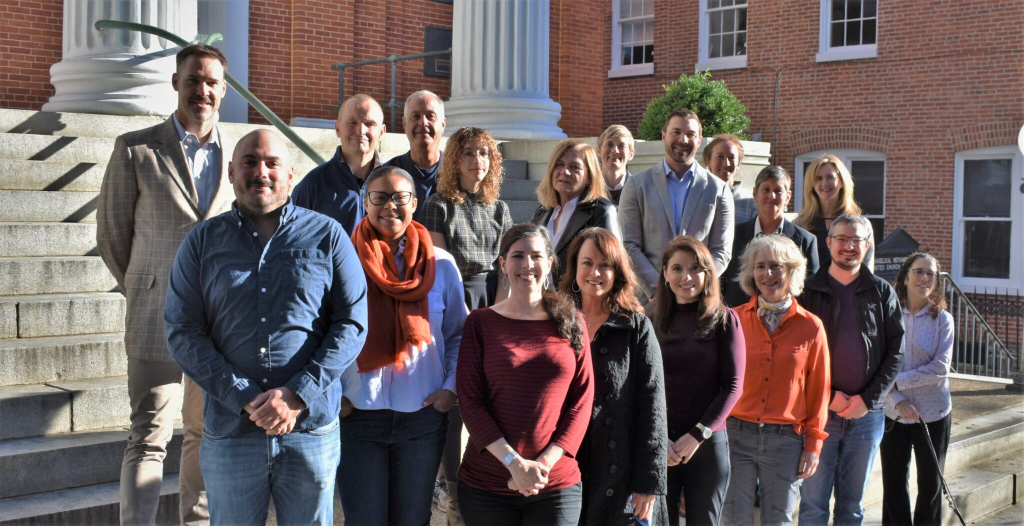 A mixed group of men and women stand on steps outside of a building. They are all smiling. 
